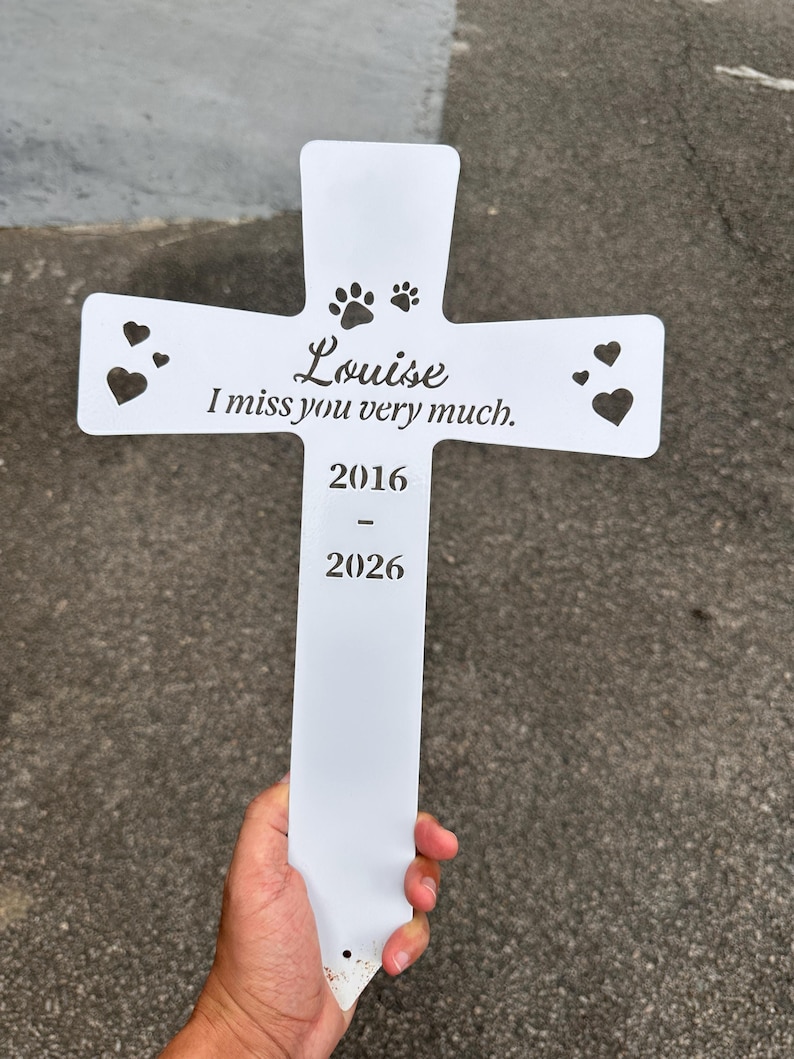 May include: A white metal cross-shaped garden stake featuring cut-out heart and paw print designs. The text reads Louise, I miss you very much, 2016, and 2026. The surface is smooth and the design is centered on the vertical and horizontal bars.