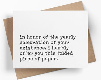May include: A white rectangular greeting card with black typewriter-style text that reads: in honor of the yearly celebration of your existence, i humbly offer you this folded piece of paper. The card is placed on top of a brown paper envelope.