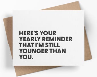 May include: A white greeting card with black text that reads: HERE'S YOUR YEARLY REMINDER THAT I'M STILL YOUNGER THAN YOU. The card is placed on top of a brown kraft paper envelope. The design is simple, featuring bold, sans-serif typography.