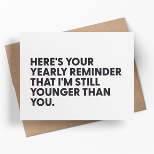 May include: A white greeting card with the text "HERE'S YOUR YEARLY REMINDER THAT I'M STILL YOUNGER THAN YOU." is placed on a brown envelope. The text is in a bold, black font.