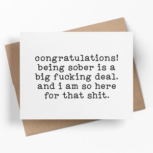 May include: A white rectangular greeting card with black typewriter-style text that reads: congratulations! being sober is a big fucking deal. and i am so here for that shit. The card is positioned on top of a brown kraft paper envelope.