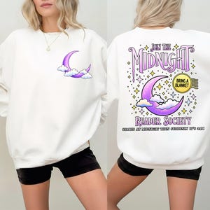 May include: White sweatshirt with a purple crescent moon and cloud design on the front. The back features the text "JOIN THE MIDNIGHT READER SOCIETY" with a moon and stars. The phrase "BRING A BLANKET" is also included.