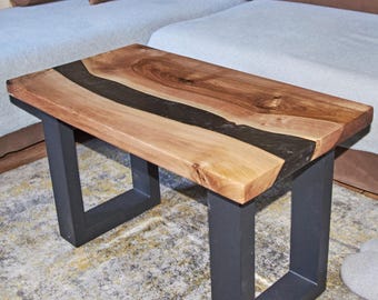 May include: A handcrafted wooden coffee table with a live edge design and a black resin inlay. The table has a rectangular top and is supported by black metal legs. The table is placed in front of a brown sectional sofa.