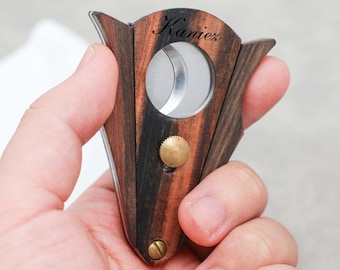 May include: A wooden cigar cutter with a stainless steel blade and brass accents. The wood has a dark, striped pattern. The word "Kanier" is engraved on the wood. The cutter is held in a hand.