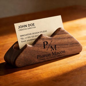 Puede incluir: Un tarjetero de madera con diseño de cordillera. Muestra una tarjeta de visita con el nombre "John Doe" y datos de contacto. El tarjetero está grabado con las iniciales "P/M" y el nombre "Pharon Macon", Manager.