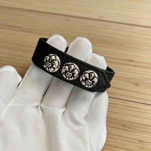 May include: Black leather bracelet with three silver fleur-de-lis studs. The bracelet is held by a gloved hand, highlighting the detailed design of the metal accents. A stylish accessory.