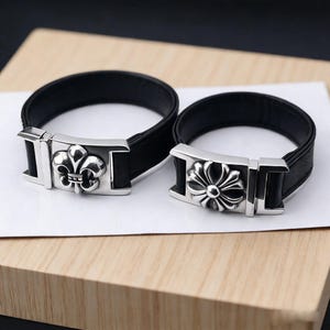 May include: Two black leather bracelets with silver clasps. One clasp has a fleur-de-lis design, the other a stylized flower. The bracelets are shown on a light wooden surface.