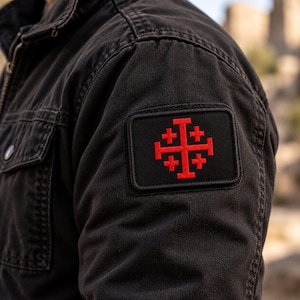 May include: A black jacket featuring a rectangular patch on the sleeve. The patch is black with a red embroidered Jerusalem cross, which consists of a large central cross surrounded by four smaller crosses in each of the four quadrants.