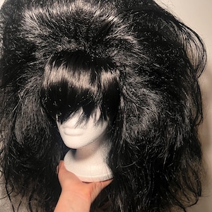 May include: A black wig with voluminous, textured hair and bangs. The wig is displayed on a white mannequin head. The wig is held by a hand with a tattoo that says "Y OUT".