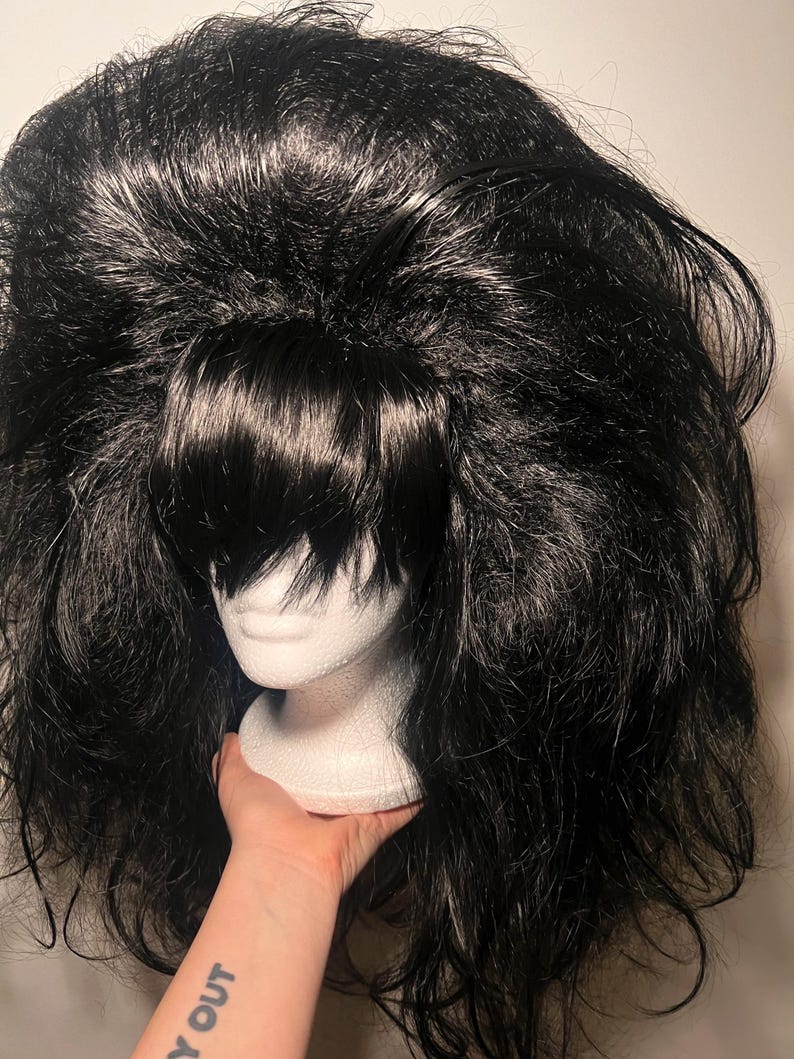 May include: A black wig with voluminous, textured hair and bangs. The wig is displayed on a white mannequin head. The wig is held by a hand with a tattoo that says "Y OUT".