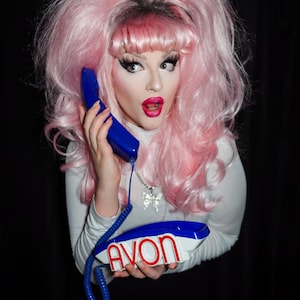 May include: A person with pink hair and makeup holds a blue and white telephone with the word "AVON" on it. The person is wearing a white turtleneck and a butterfly necklace. The background is black.