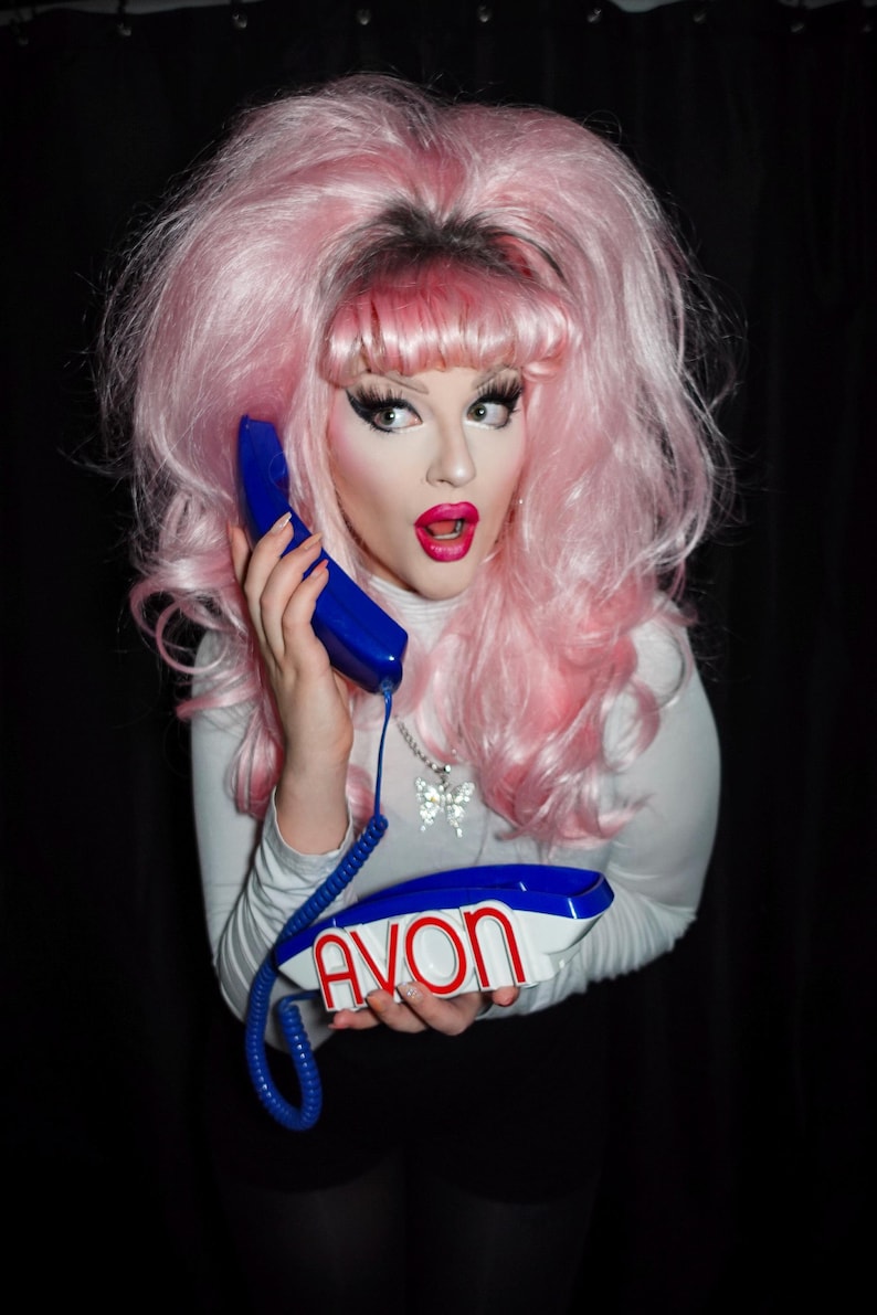 May include: A person with pink hair and makeup holds a blue and white telephone with the word "AVON" on it. The person is wearing a white turtleneck and a butterfly necklace. The background is black.