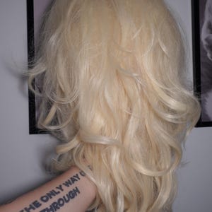 K&ouml;nnte beinhalten: Eine lange, wellige, blonde Per&uuml;cke mit nat&uuml;rlicher Textur. Die Per&uuml;cke wird vor einer Wand mit gerahmten Bildern gehalten. Der Arm, der die Per&uuml;cke h&auml;lt, hat ein Tattoo mit der Aufschrift "THE ONLY WAY IS THROUGH."