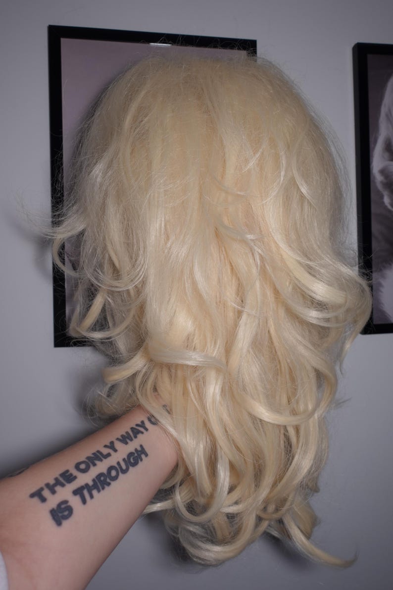 K&ouml;nnte beinhalten: Eine lange, wellige, blonde Per&uuml;cke mit nat&uuml;rlicher Textur. Die Per&uuml;cke wird vor einer Wand mit gerahmten Bildern gehalten. Der Arm, der die Per&uuml;cke h&auml;lt, hat ein Tattoo mit der Aufschrift "THE ONLY WAY IS THROUGH."