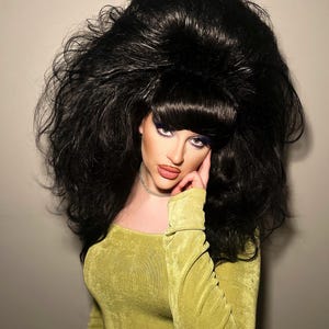 May include: A person wearing a voluminous black wig with a bouffant style. The person is wearing a long-sleeved, olive green dress and a silver choker necklace. The person is looking at the camera.