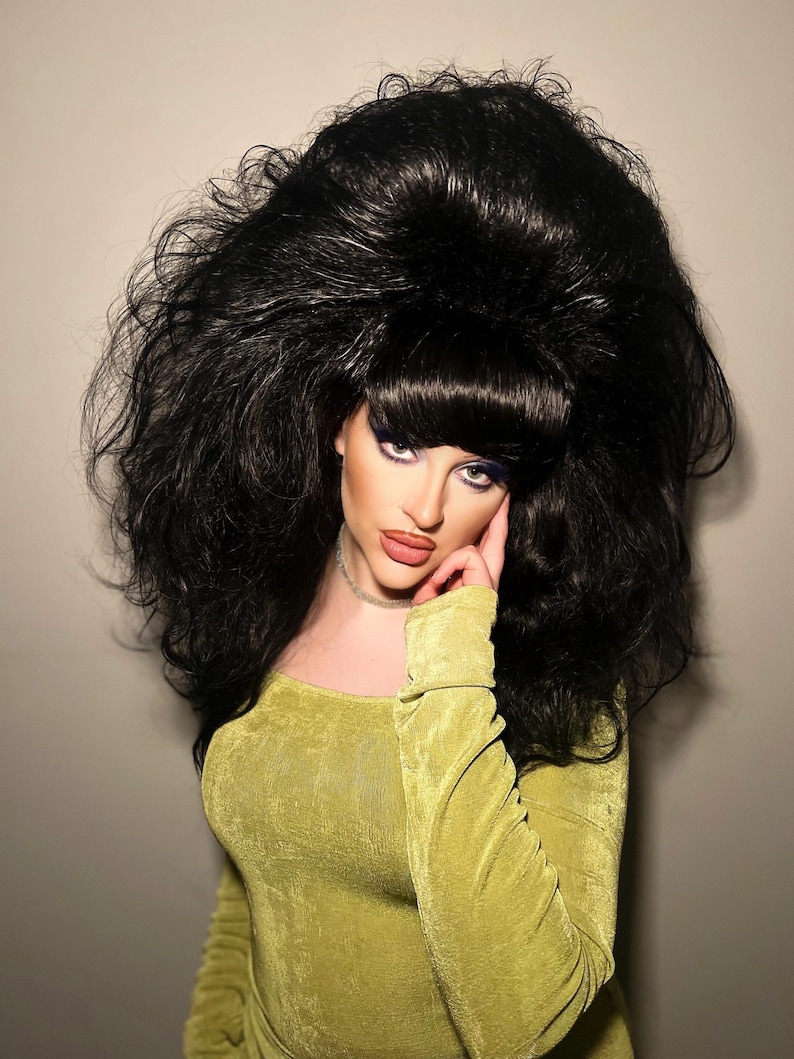 May include: A person wearing a voluminous black wig with a bouffant style. The person is wearing a long-sleeved, olive green dress and a silver choker necklace. The person is looking at the camera.