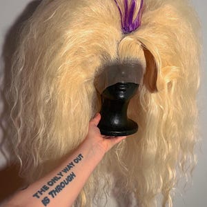 May include: A long, wavy blonde wig with a dark lace front and a purple accent. The wig is styled on a black wig stand. The arm holding the wig stand has a tattoo that reads "THE ONLY WAY OUT IS THROUGH."