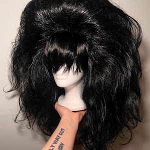 May include: A voluminous black wig with a short, blunt fringe, styled with lots of volume. The wig is displayed on a white mannequin head. The arm holding the head has a tattoo that reads "THE ONLY WAY OUT IS THROUGH."