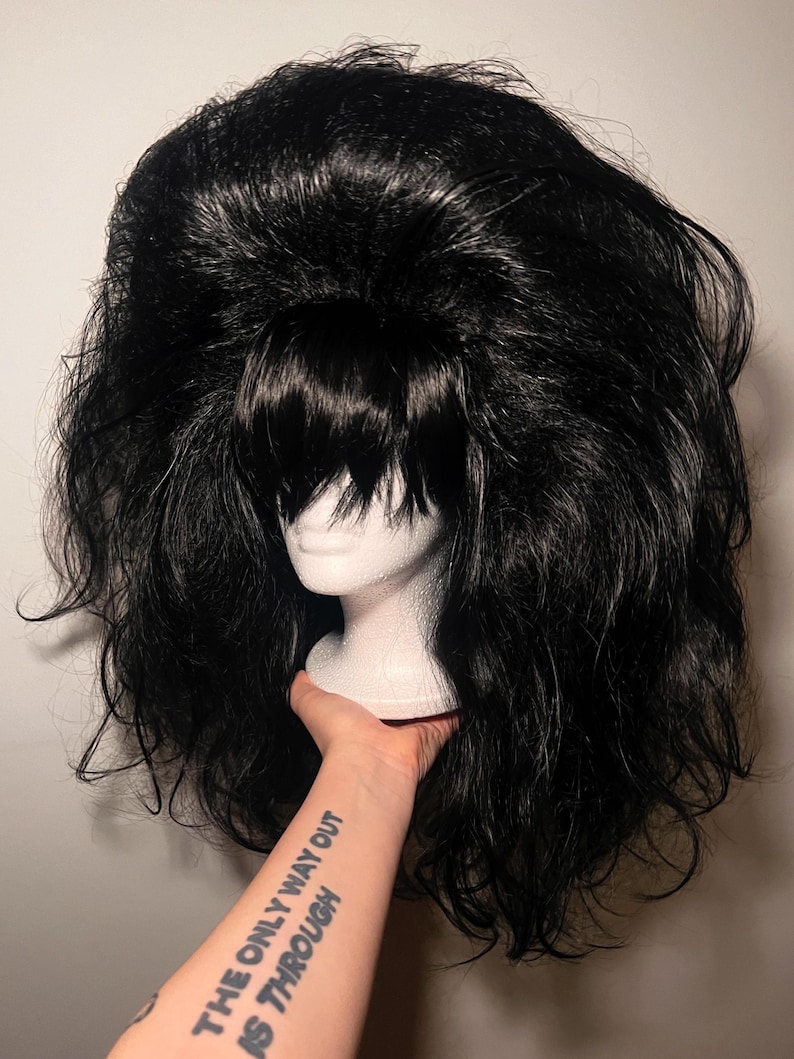 May include: A voluminous black wig with a short, blunt fringe, styled with lots of volume. The wig is displayed on a white mannequin head. The arm holding the head has a tattoo that reads "THE ONLY WAY OUT IS THROUGH."
