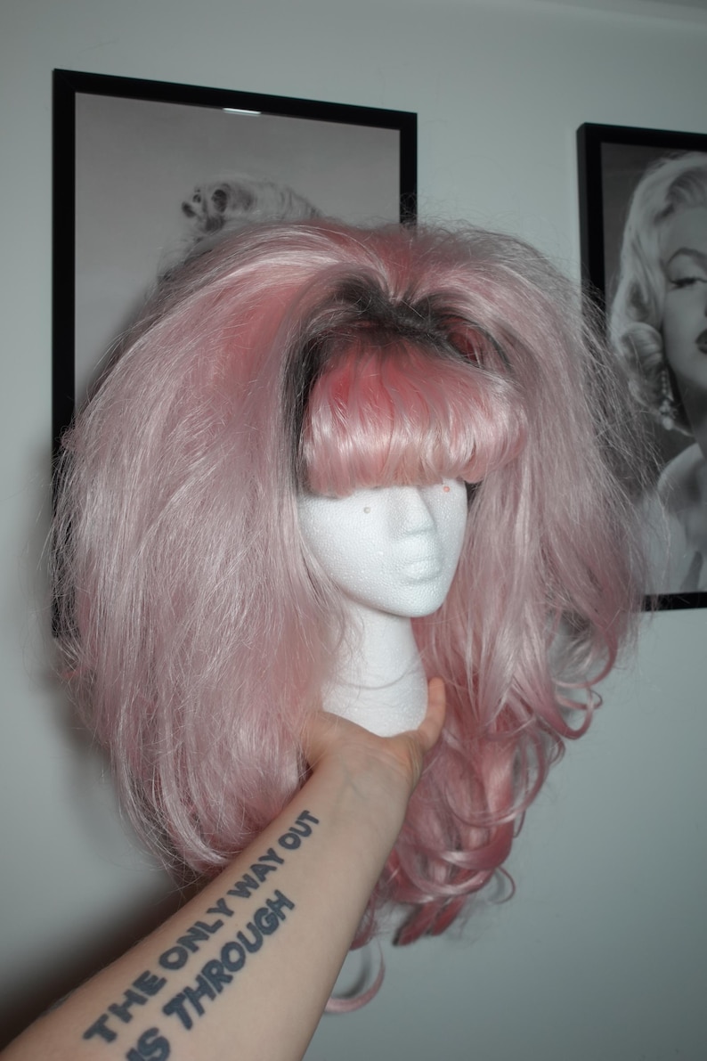 May include: A pink wig with bangs is displayed on a styrofoam head. The wig has long, voluminous hair with darker roots. The arm holding the head has a tattoo that reads "THE ONLY WAY OUT IS THROUGH".