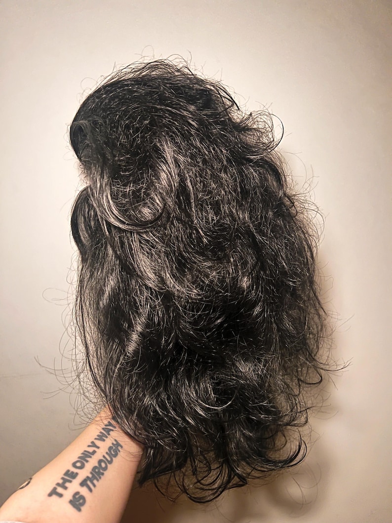 May include: A dark gray wig with wavy, layered hair. The wig has a natural look with some lighter gray strands mixed in. The hair is styled with volume and texture, creating a full and realistic appearance.