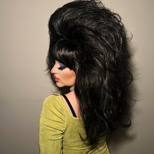May include: A person with a large, voluminous black wig and a lime green dress. The wig is styled with a high bouffant and bangs. The dress has long sleeves and a square neckline. The person is facing away from the camera.