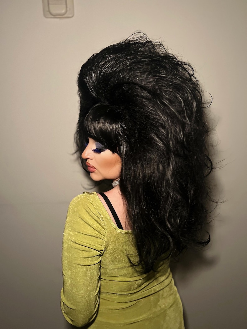 May include: A person with a large, voluminous black wig and a lime green dress. The wig is styled with a high bouffant and bangs. The dress has long sleeves and a square neckline. The person is facing away from the camera.