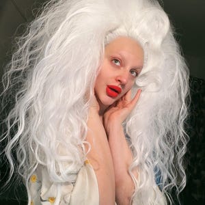 May include: A person wearing a large, voluminous white wig with long, wavy strands. The person has pale skin, red lipstick, and is wearing a cream-colored garment with a floral pattern. The person is looking upwards with a thoughtful expression.