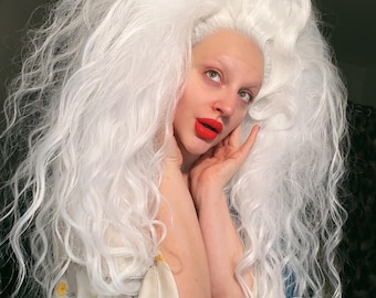 May include: A person wearing a large, voluminous white wig with long, wavy strands. The person has pale skin, red lipstick, and is wearing a cream-colored garment with a floral pattern. The person is looking upwards with a thoughtful expression.