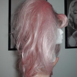 May include: A light pink wig styled with voluminous waves and a side-swept bang, displayed on a white mannequin head. The wig is held up by a hand with a tattoo that says "LY WAY OUT GH".