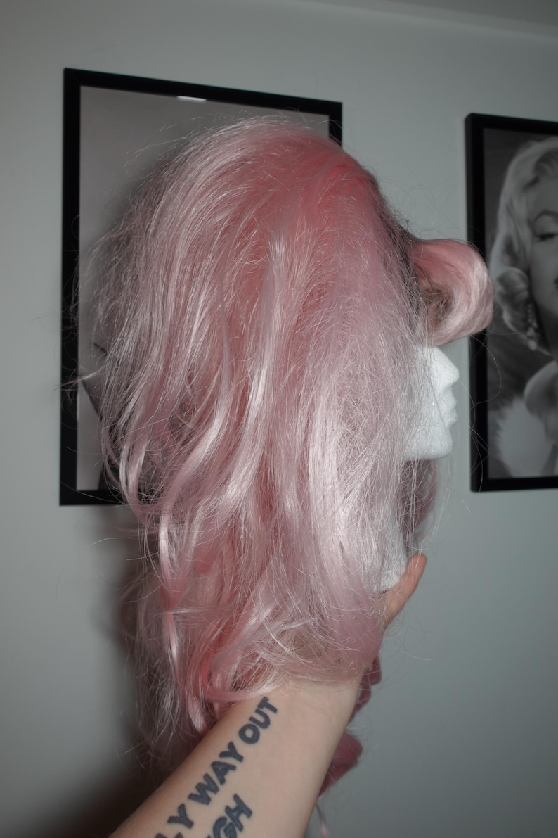 May include: A light pink wig styled with voluminous waves and a side-swept bang, displayed on a white mannequin head. The wig is held up by a hand with a tattoo that says "LY WAY OUT GH".