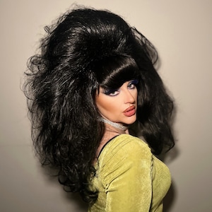 May include: A person with dramatic makeup and a large, voluminous black wig. The person is wearing a chartreuse green dress and a silver choker necklace. The background is a plain, light-colored wall.