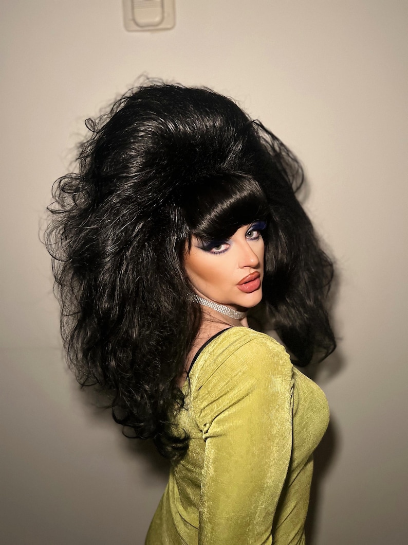 May include: A person with dramatic makeup and a large, voluminous black wig. The person is wearing a chartreuse green dress and a silver choker necklace. The background is a plain, light-colored wall.
