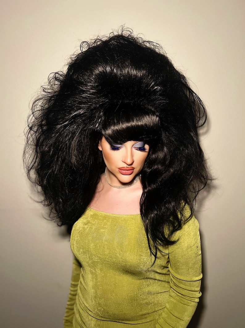 May include: A person wearing a large, voluminous black wig with a bouffant style. The person is wearing a long-sleeved, olive green dress and has dramatic makeup with blue eyeshadow and red lipstick.