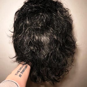 May include: A dark, curly wig with strands of gray. The wig is held by a person's arm, with a tattoo that reads "THE ONLY WAY IS THROUGH". The wig appears to be styled with a natural, tousled look.