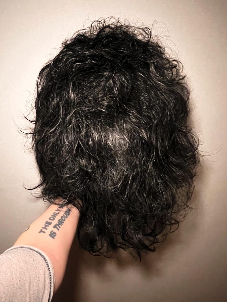 May include: A dark, curly wig with strands of gray. The wig is held by a person's arm, with a tattoo that reads "THE ONLY WAY IS THROUGH". The wig appears to be styled with a natural, tousled look.