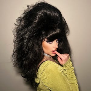 May include: A person with dramatic makeup and a large, voluminous black wig. They are wearing a chartreuse green long-sleeved dress and a silver choker. The person is touching their lip with a finger.