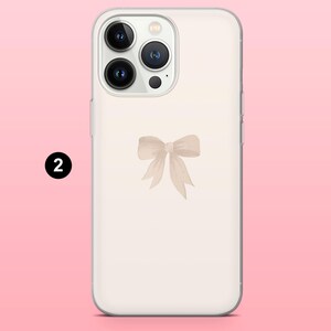 May include: A clear phone case featuring a light beige bow design. The bow is centered on the case and has a watercolor effect. The phone case has a clear border and a pink background.