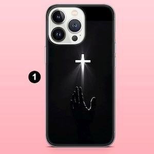 May include: Black phone case featuring a white cross with light rays emanating from it, above a silhouetted hand reaching upwards. The case has a clear border and a black background.