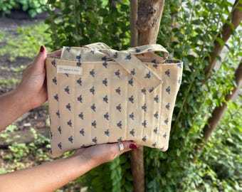 May include: A beige tote bag with a repeating pattern of blue bees. The bag has a matching bow and handles. The text "handmade by fanny" is on a small white tag. The bag is held by two hands.