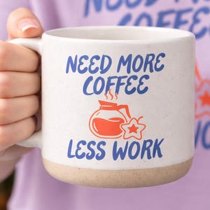 Puede incluir: Taza de café de cerámica color crema con el texto "NEED MORE COFFEE LESS WORK" en azul y naranja. La taza tiene una base beige y un asa blanca. La imagen también muestra a una persona con una camiseta morada.