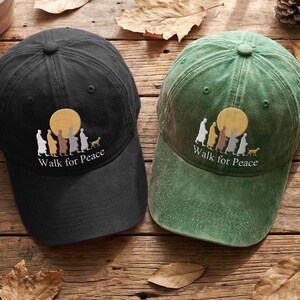 May include: Two baseball caps, one black and one green, are shown on a wooden surface. Each cap has an embroidered design of figures walking towards a yellow sun, with the text "Walk for Peace" below. The caps are surrounded by leaves and a small succulent plant.
