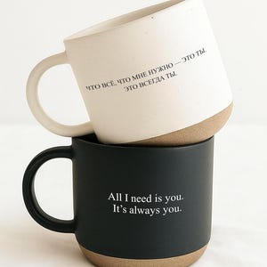 May include: Two ceramic mugs, one stacked atop the other. The top mug is white with a tan base and a curved handle. The bottom mug is black with a tan base and a curved handle. Text is printed on both mugs.