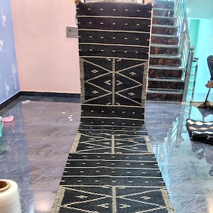 Puede incluir: Una alfombra pasillera larga y rectangular de color negro con un patrón repetitivo de formas geométricas de diamante blancas y líneas horizontales discontinuas. La alfombra tiene un tejido texturizado y flecos cortos claros.