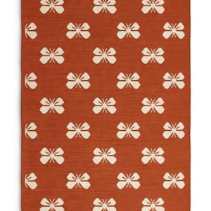 May include: A rectangular area rug featuring a burnt orange background with a repeating pattern of off-white butterfly motifs. The rug is finished with short, dark gray fringe along the top and bottom edges, creating a textured border.