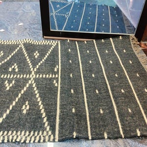 Puede incluir: Una alfombra gris y blanca estampada con un diseño geométrico en el lado izquierdo y rayas verticales con puntos en el derecho. La alfombra tiene un borde con flecos y está hecha de material tejido. La alfombra se muestra sobre una superficie gris.