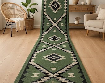 May include: A long, green runner rug with a geometric pattern in black and cream. The rug features a diamond and zigzag design, with a fringe at the bottom. The rug is displayed indoors on a hardwood floor.