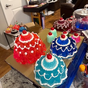 Op de afbeelding: Een verzameling kleurrijke, koepelvormige decoratieve items die lijken op gelatinedesserts. De items zijn in tinten rood, blauw, teal en bruin, bekroond met een kers. Witte, frosting-achtige details sieren de randen.