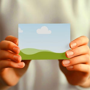 May include: A rectangular card featuring a landscape design with a blue sky, white clouds, and green rolling hills. The card is held by two hands, with the focus on the card's design.