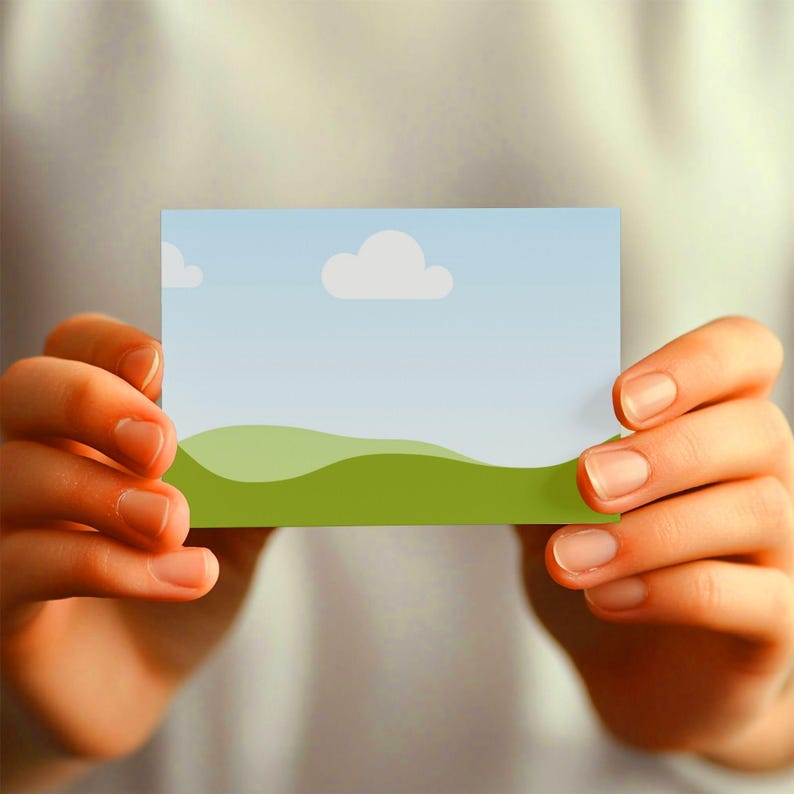 May include: A rectangular card featuring a landscape design with a blue sky, white clouds, and green rolling hills. The card is held by two hands, with the focus on the card's design.